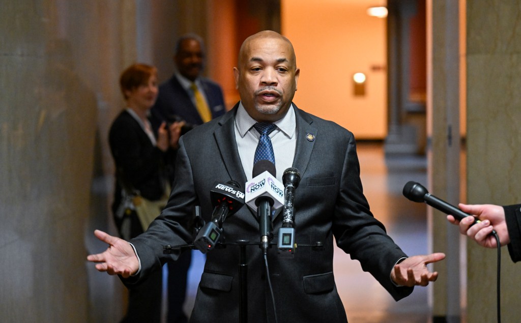 Carl Heastie speaking in a hallway inside the Capitol with his arms outstretched as he speaks in front of blurry people