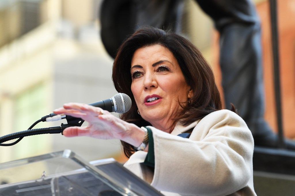 Kathy Hochul speaking at a microphone in front of a blurry background with her left arm extended.