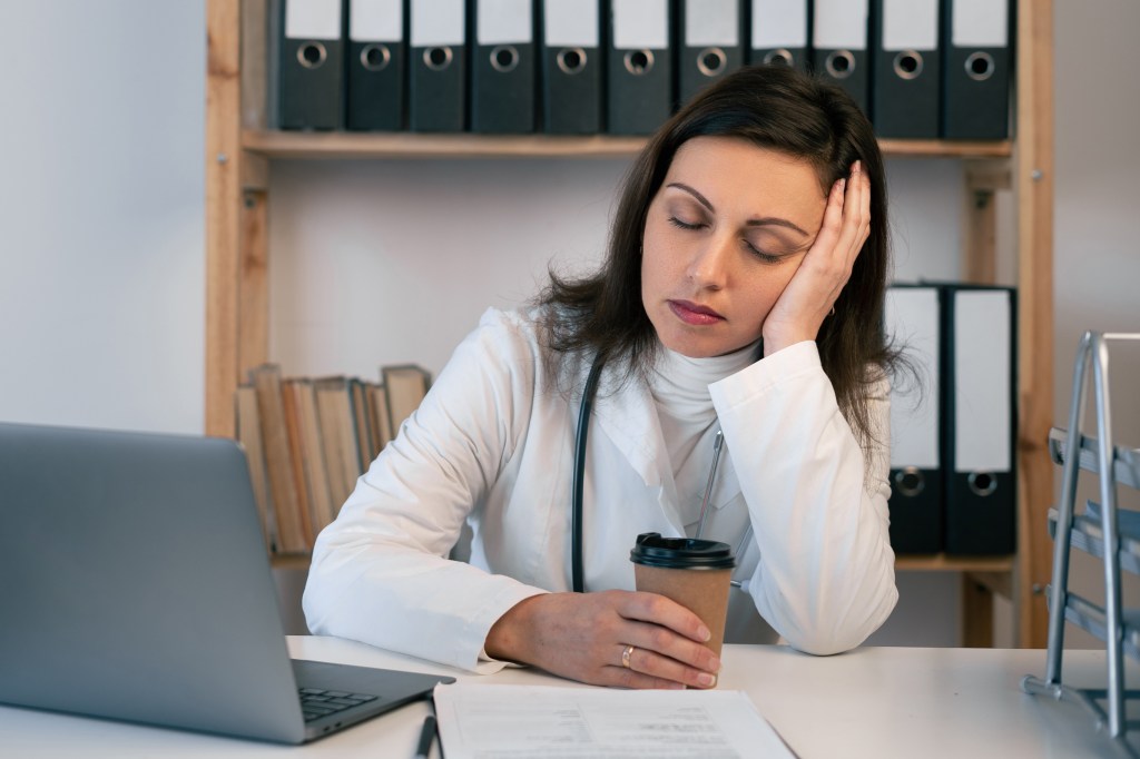 Tired female doctor trying to stay awake with glass of coffee while working at her office. Overworked and overtime concept. Copy space. Banner