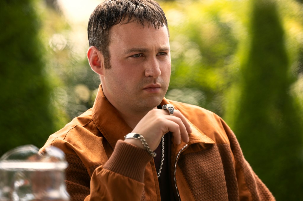 Emory Cohen looking serious sitting in a chair outside. 