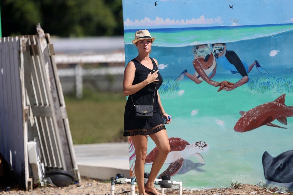 Jasmine Hartin in a black dress and straw hat walks by a wall with a painted mural.