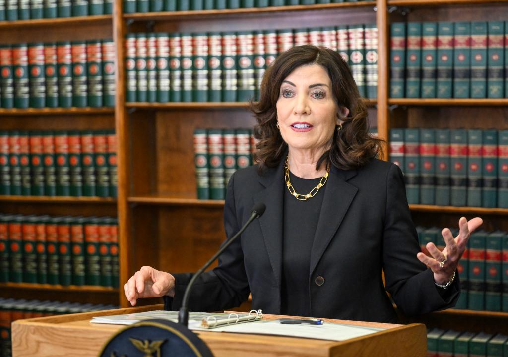 Kathy Hochul speaking at a wooden podium with her left hand raised as she speaks in front of bookshelves filled with heavy legal books. 