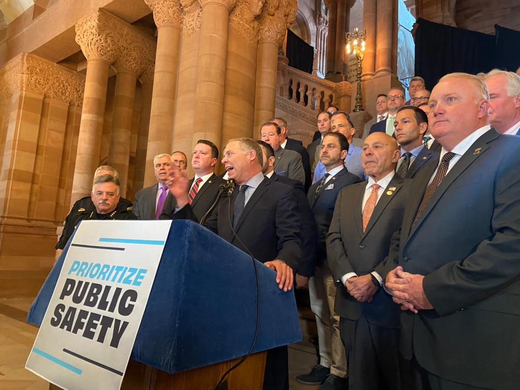 Will Barclay speaking at a podium with rows of white Republican men standing on a brownstone staircase inside the state Capitol 