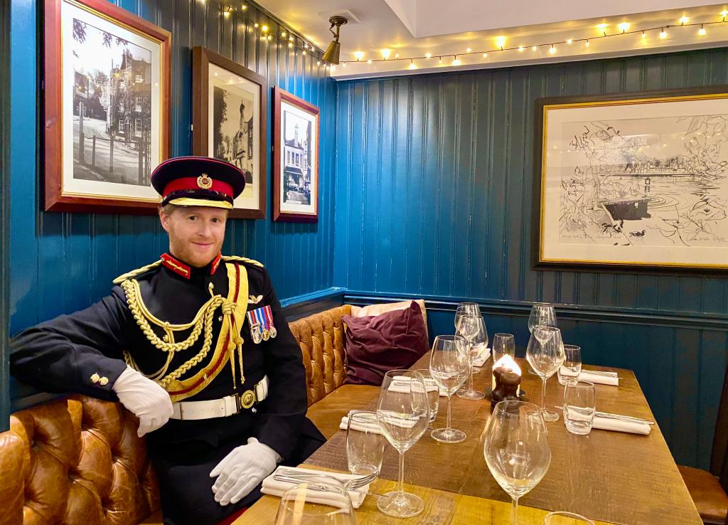 Man dressed up as Prince Harry sitting at table with glass wear.