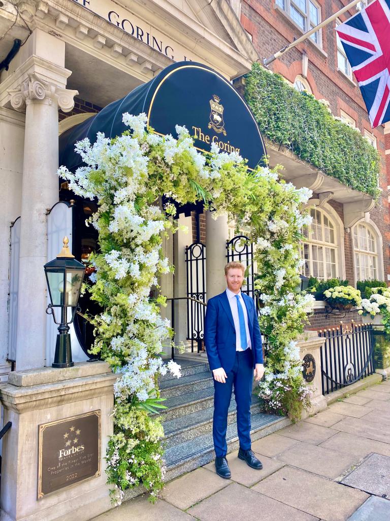 Red-haired man in blue suit in front of flowered entrance.