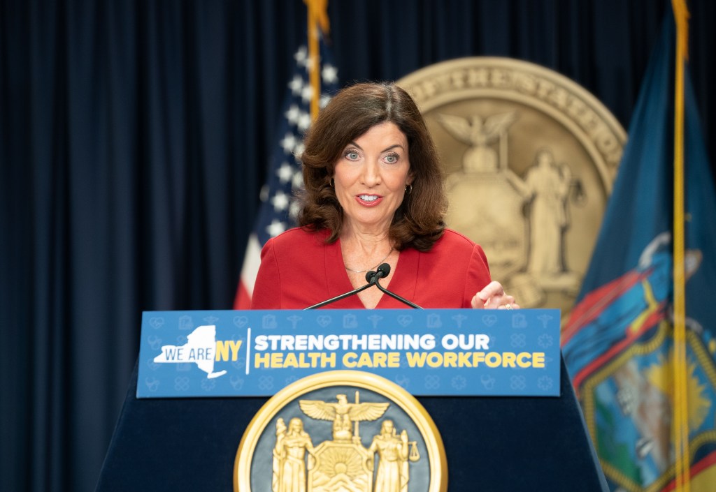 Kathy Hochul speaking at a podium inside her NYC office with a big state seal and flags behind her