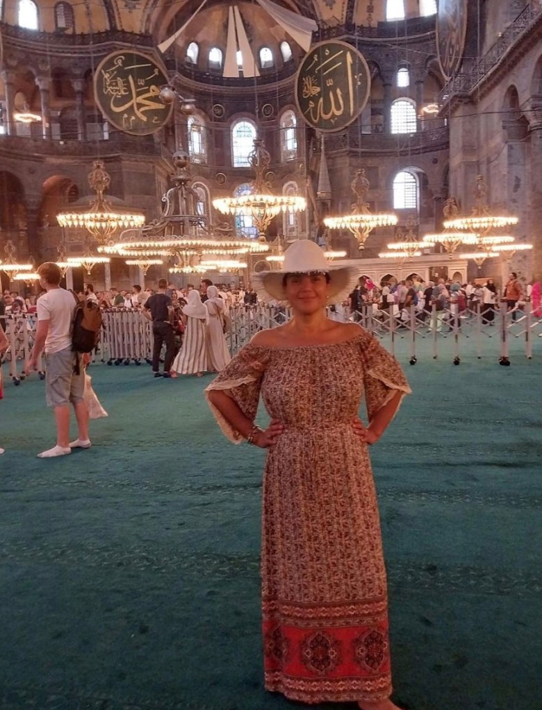 Woman big white hat and floral red dress hands on hips.