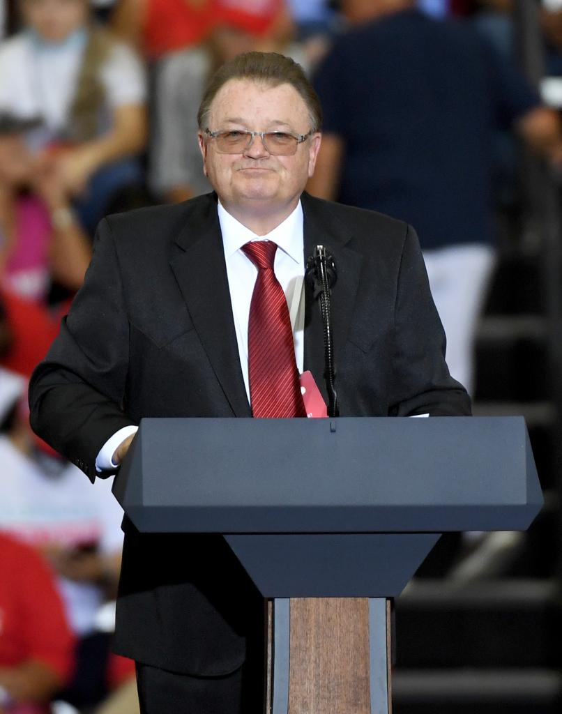 A man in a suit and tie standing at a podium speaking in front of a blue background.