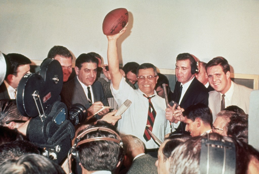 Green Bay Packers coach Vince Lombardi holds a football overhead surrounded by reporters. 