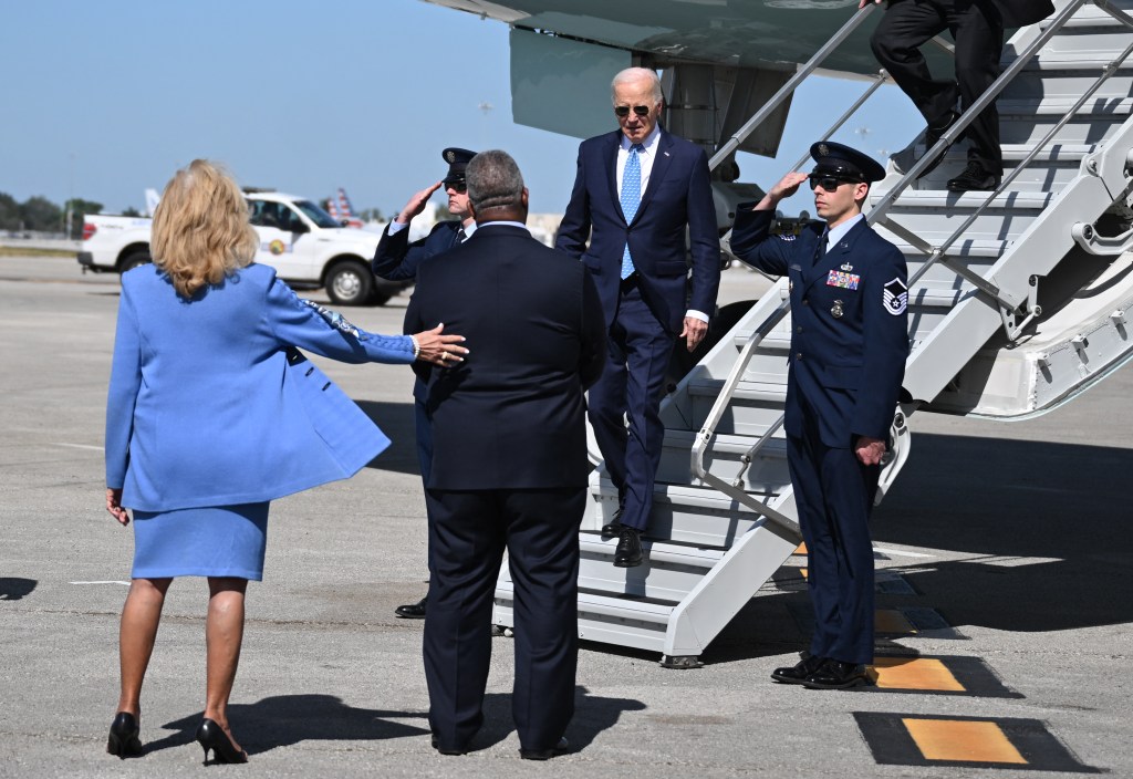 US President Joe Biden walks down the steps of Air Force One as an Airman salutes and two people wait to greet him.