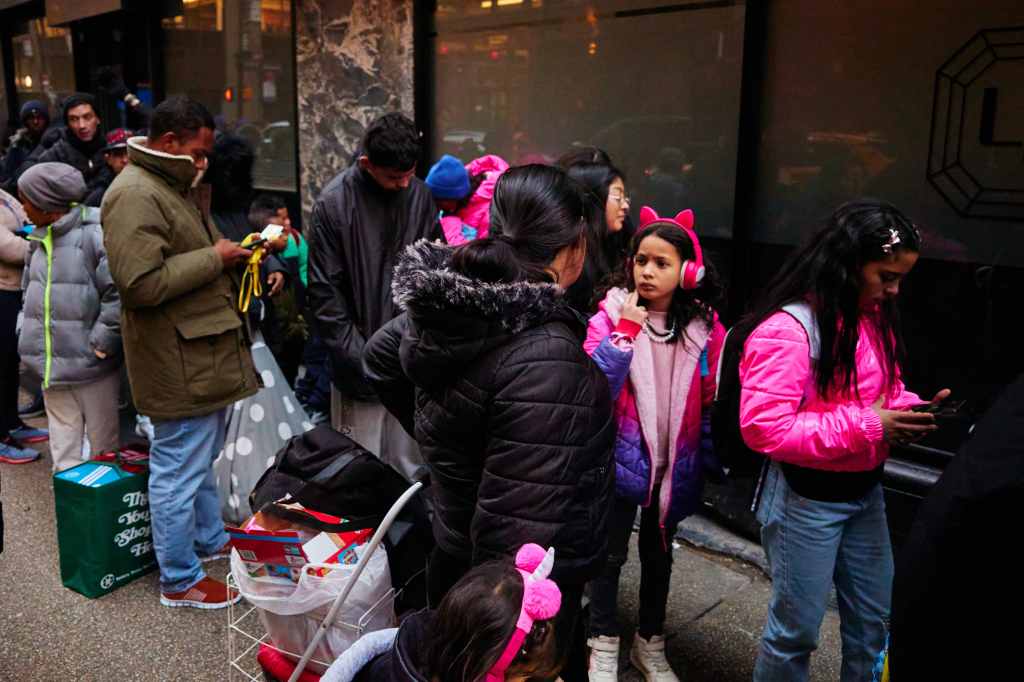 A group of migrants, including a young girl in a pink and blue jacket wearing pink headphones, line up in front of a building in New York City