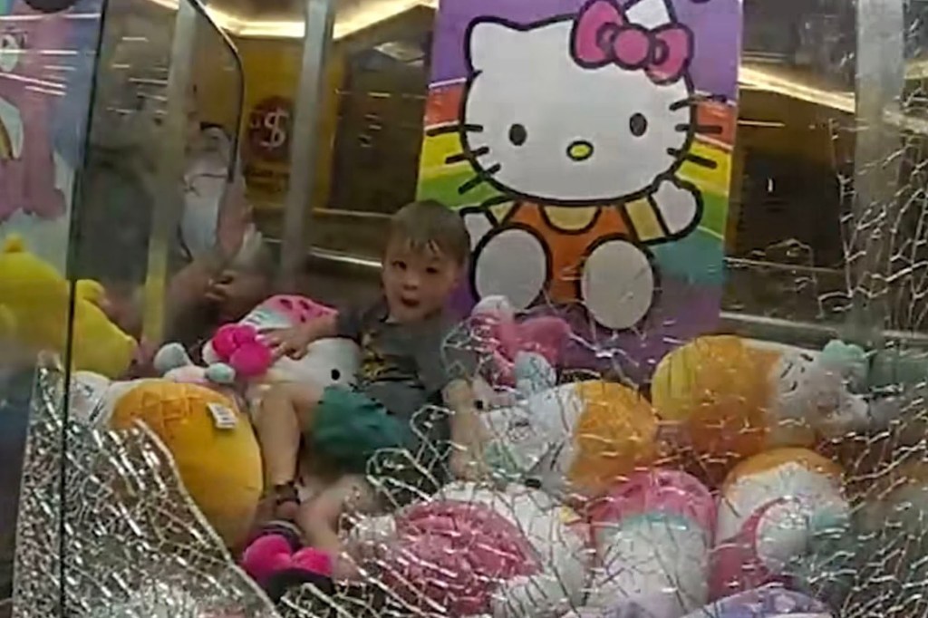 A boy sitting on a pile of stuffed toys inside a claw machine at a shopping center.