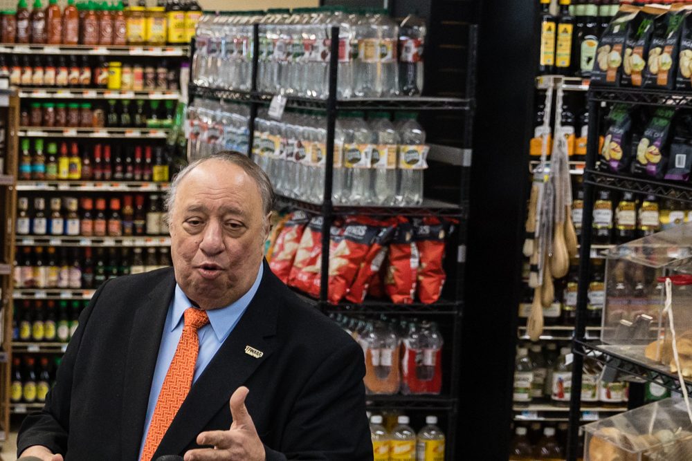 John Catsimatidis standing in front of supermarket shelves containing water, chips and sauces