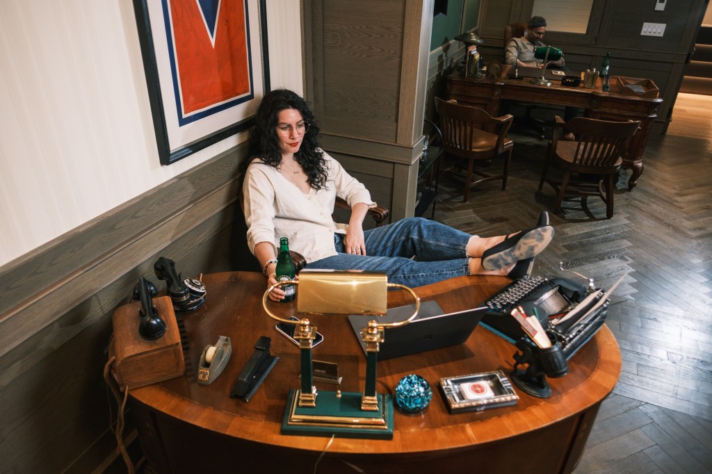 A woman sits at a 1940s detective's desk, her feet up on the desk. 