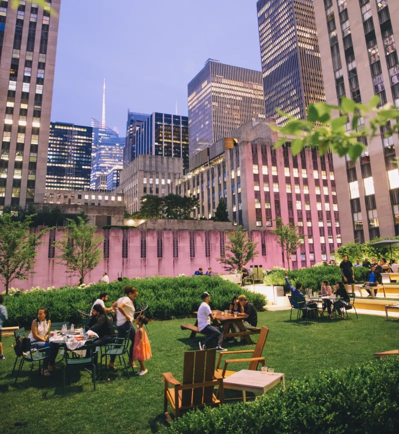 Somewhat cropped shot of Radio Park atop Rockefeller Center. People walk around or sit and socialize                at the three tables within the shot. 