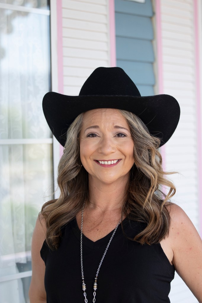 A woman smiles at the camera in her black tank top and black cowboy hat.