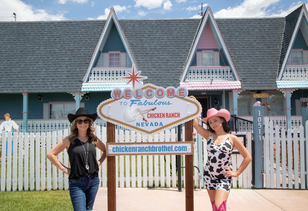 Woman in black tank top, jeans, sunglasses and a black cowboy hat stands with her hands on her hips on the left side of a sign that says, "Welcome to Fabulous Chicken Ranch Nevada" and chickenranchbrothel.com. On the right side of the sign is a woman in a cowprint dress, thigh-high pink cowboy boots and a pink cowboy hat with her hand on the sign.