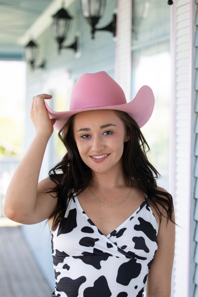 Woman in a cow dress and pink cowboy hat smiles at the camera with her hand on the hat.