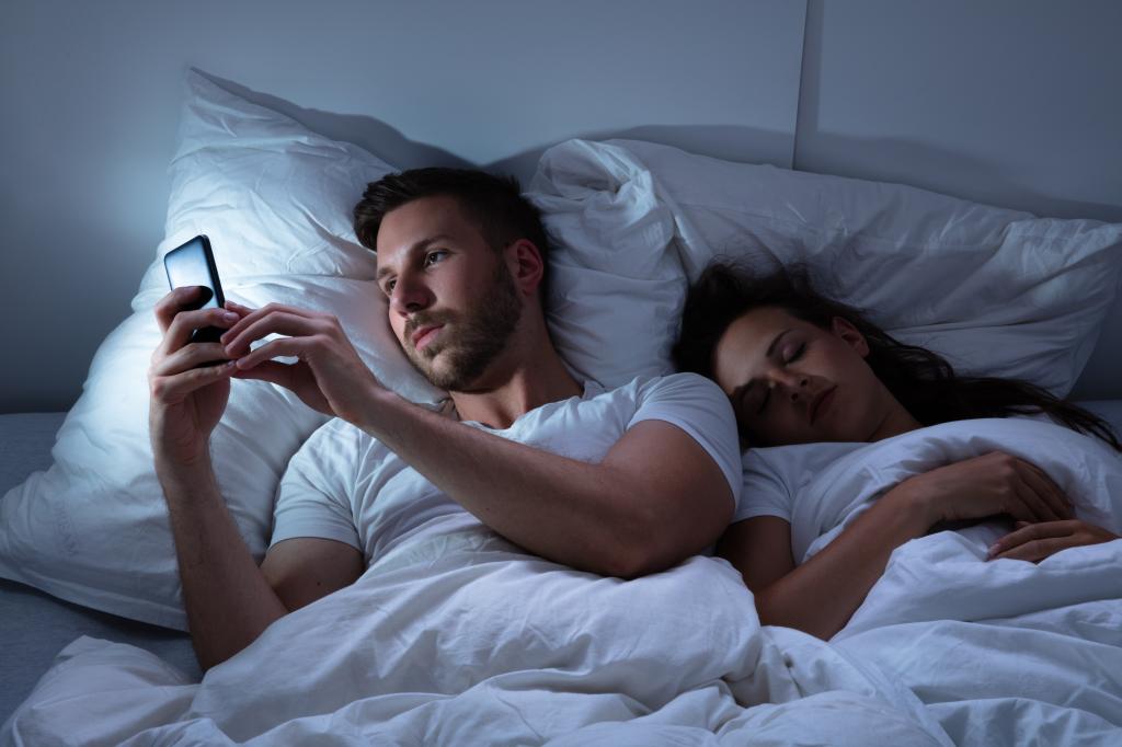 Young man using cellphone in bed at night with his sleeping partner nearby