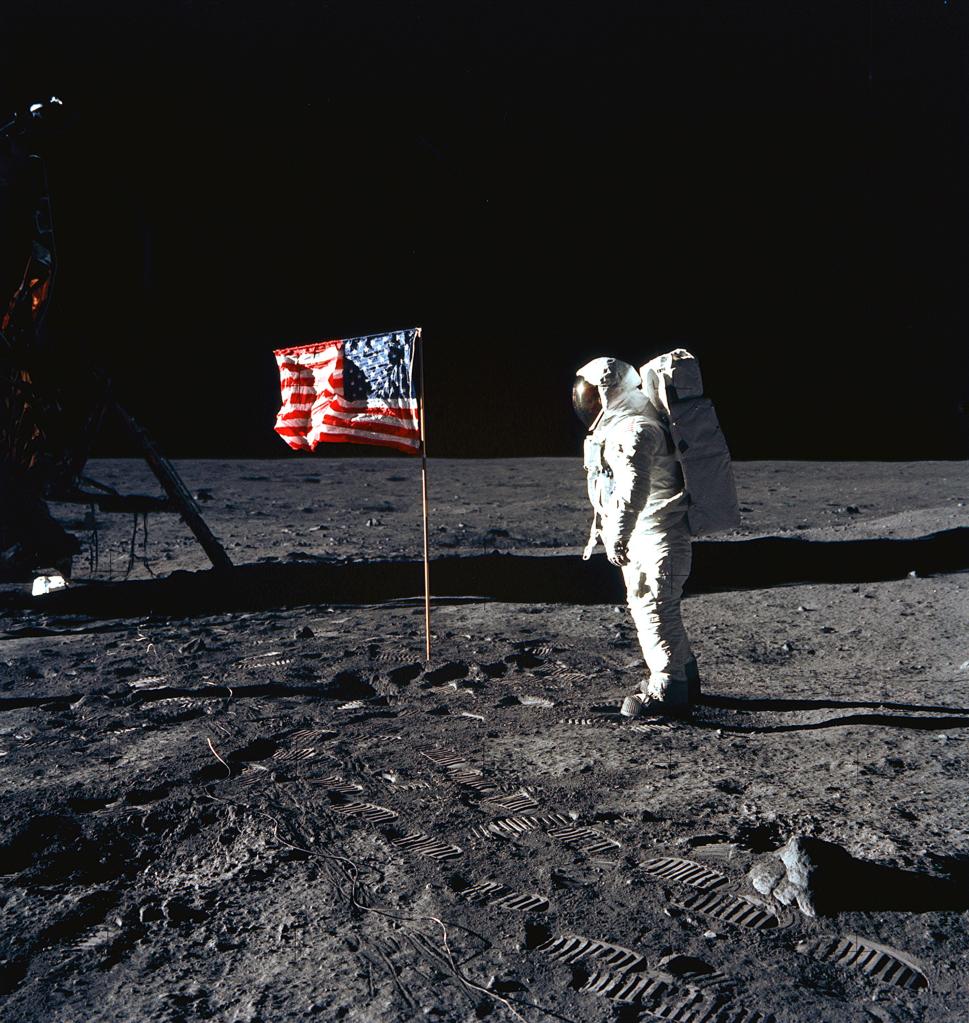 Astronaut Edwin E. Aldrin standing next to the United States flag deployed on the lunar surface, with lunar module and astronaut footprints visible, during Apollo 11 mission