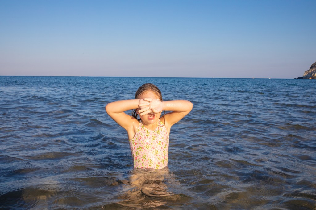 little girl with swimsuit in the water of Mediterranean sea covering her eyes with hand