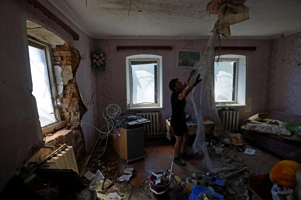 A man cleaning up his mangled home, which was damaged by recent Russian shelling. The windows are covered with plastic, exposed bricks are visible in one window and debris covers the floor, while the man deals with materials falling from the ceiling.