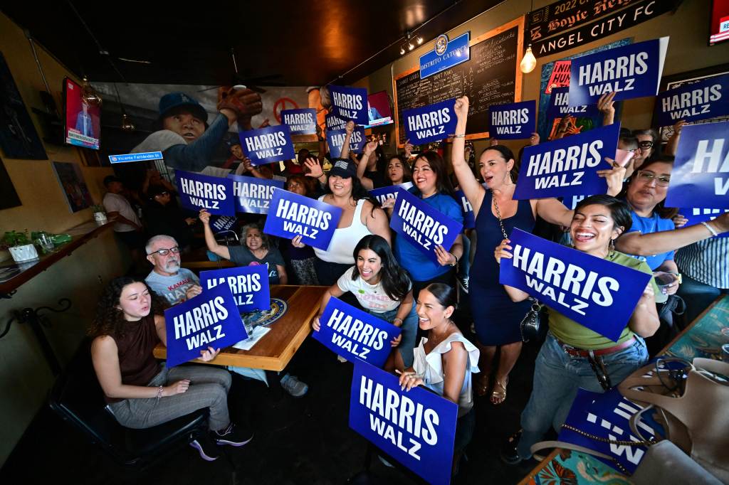 Supporters of the Latinos for Harris-Walz campaign, including Chrissie Fit and Sandy Koufax, watching Kamala Harris delivering her acceptance speech on screen at a party.