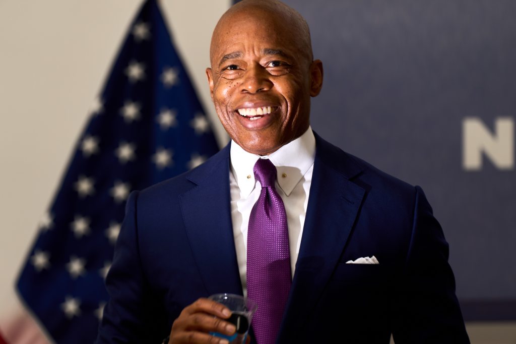 New York City Mayor Eric Adams giving a speech at a news conference in Queens Museum, smiling in a suit