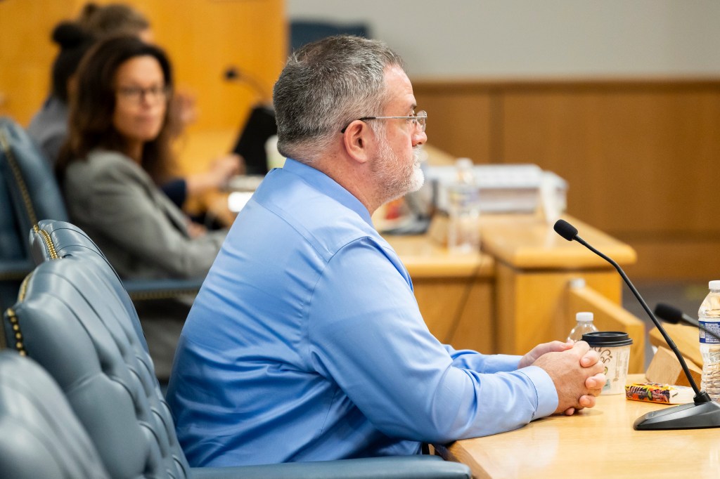 Matthew McCoy, a former OceanGate employee, listening to questions at the Coast Guard investigatory hearing about the Titanic tourist sub incident, seen from side wearing glasses and blue dress shirt, mic in front of him.