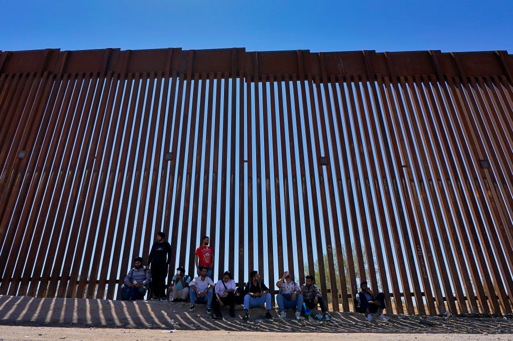 A group of people, claiming to be from India, sitting in the shade of the US-Mexico border wall waiting for Border Patrol pickup in Arizona, 2023