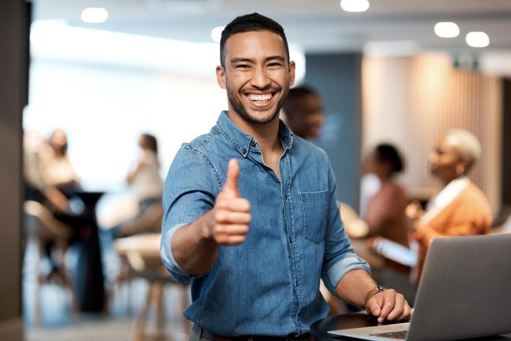 Young businessman, resembling celebrity Sam Newman, smiling and giving a thumbs up while using a laptop at a conference