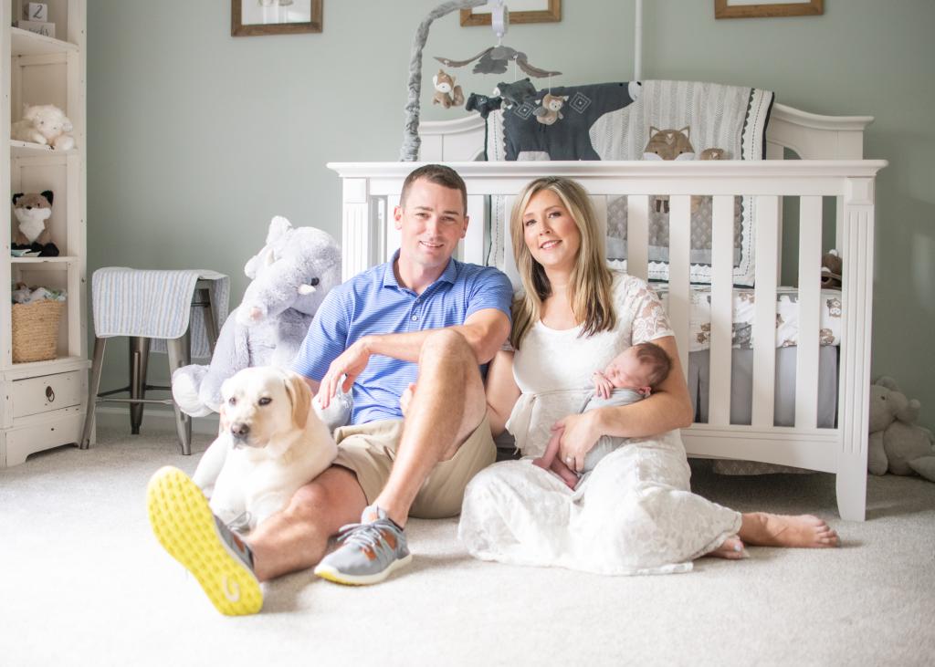 John Willard and Lauren Sisler sit on the floor of their son's nursery, in front of a crib. Lauren holds a baby and John sits next to a dog 