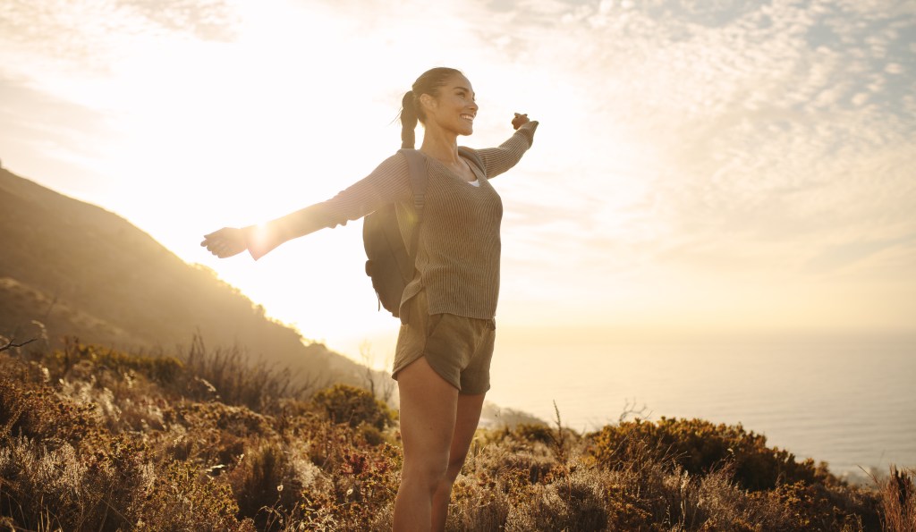 Woman hiker with backpack joyfully standing on a country path, hands outstretched, in bright sunlight