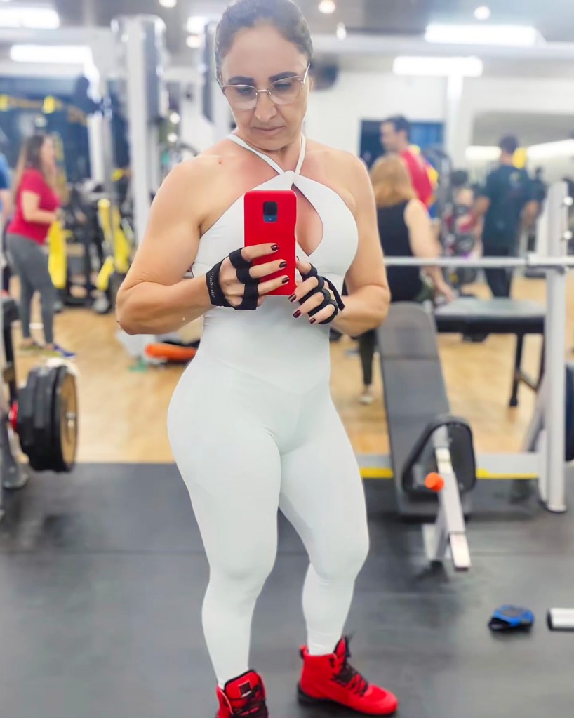 Claudia Oliveira in a one-piece workout bodysuit at the gym, taking a selfie in the mirror.