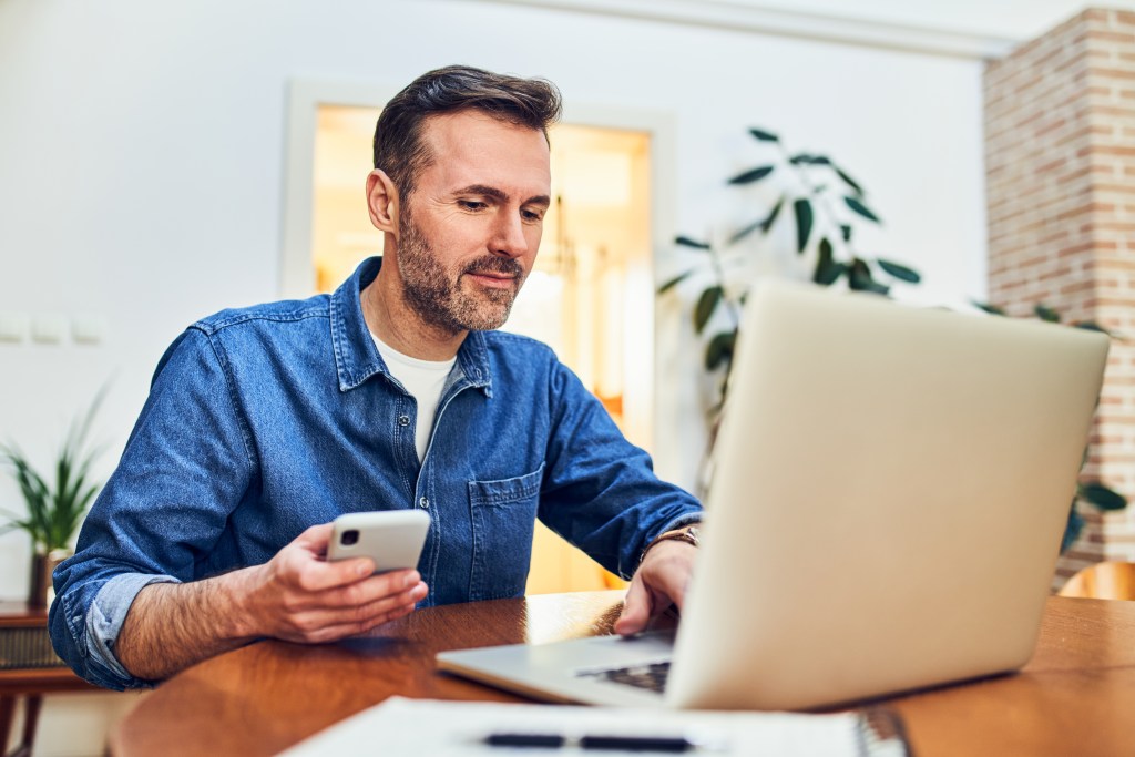 Adult man at home, sitting at a desk, making an online payment using two-factor authentication on his mobile phone