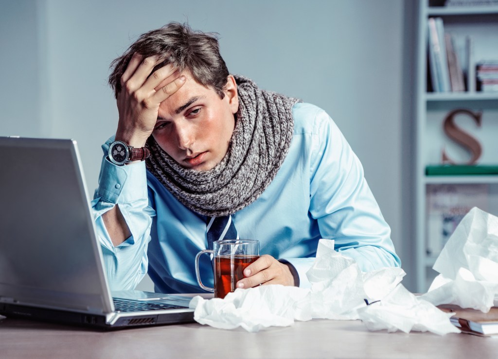 Young man feeling sick at the office, wearing a scarf and working on a laptop