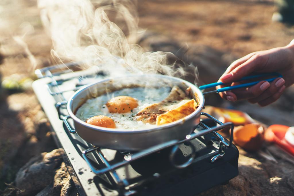 Person cooking fried eggs on a metal gas stove during an outdoor camping trip