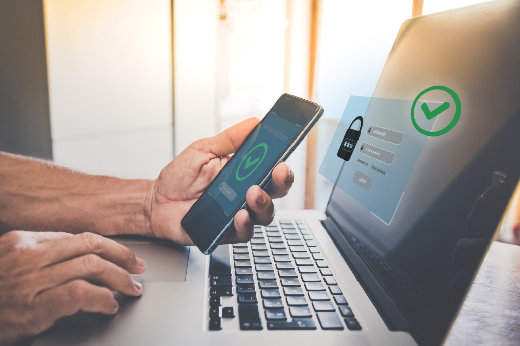 A person holding a smartphone and using a laptop for two-factor authentication, ensuring online account security