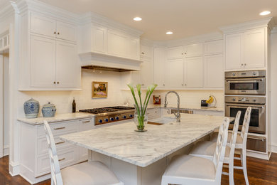 Example of a mid-sized classic l-shaped medium tone wood floor and brown floor enclosed kitchen design in Raleigh with a farmhouse sink, beaded inset cabinets, white cabinets, marble countertops, white backsplash, ceramic backsplash, stainless steel appliances, an island and white countertops