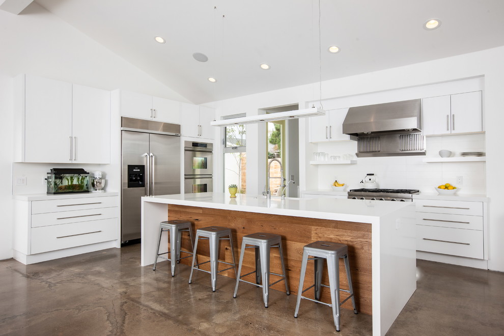 Example of a mid-sized trendy l-shaped concrete floor and brown floor kitchen design in Orange County with white cabinets, quartz countertops, stainless steel appliances, flat-panel cabinets, white backsplash and an island