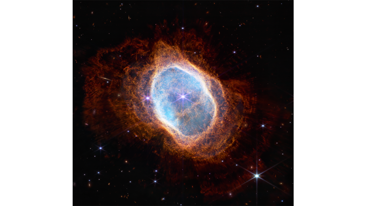 Colorful image of near-infrared light from a glowing cloud with a distorted ring-like shape, illuminated from within by a bright central star. The Southern Ring Nebula is a large, semi-transparent oval that is slightly angled from top left to bottom right. A bright white star appears at the center of this image. A large transparent teal oval surrounds the central star. Several red shells surround the teal oval, extending almost to the edges of the image. The shells become a deeper red with distance from the center. The bright central star has eight diffraction spikes. Behind the gaseous teal layers are deeper orange layers that are arranged like threads in a complex weaving. The red layers, which are wavy overall, look like they have very thin straight lines piercing through them, which are holes where light from a central star is traveling. The background of the image is black and speckled with tiny bright stars and distant galaxies.