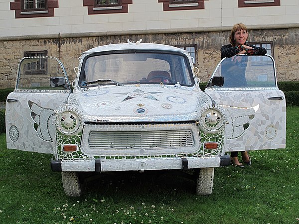 Ein komplett behäkelter Trabbi von vorne mit offenen Türen und der herstellenden Künstlerin Anne Schwegmann-Fielding