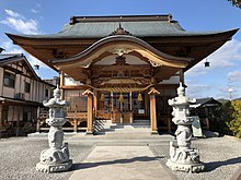 Frontalansicht eines kleinen Shintō-Schreins. Auf dem Weg zum Schrein befindet sich links und rechts eine weiße Steinsäule, die jeweils mit sie umwindenden Schlangen verziert ist.