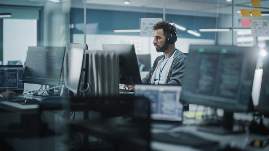 Diverse Office: White IT Programmer Working on Computer. Male Specialist Creating Innovative Software. Engineer Developing App, Program, Video Game. Wearing Headphones Listening Podcast, Music