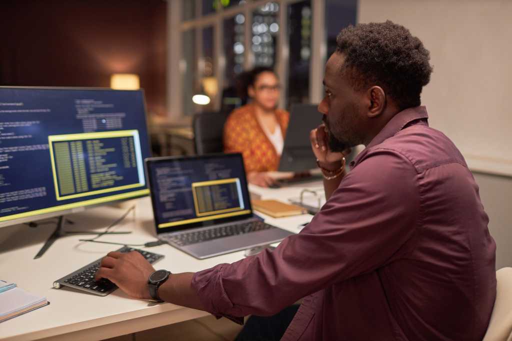 African American developer writing computer codes on computer while working at his workplace in office till late night