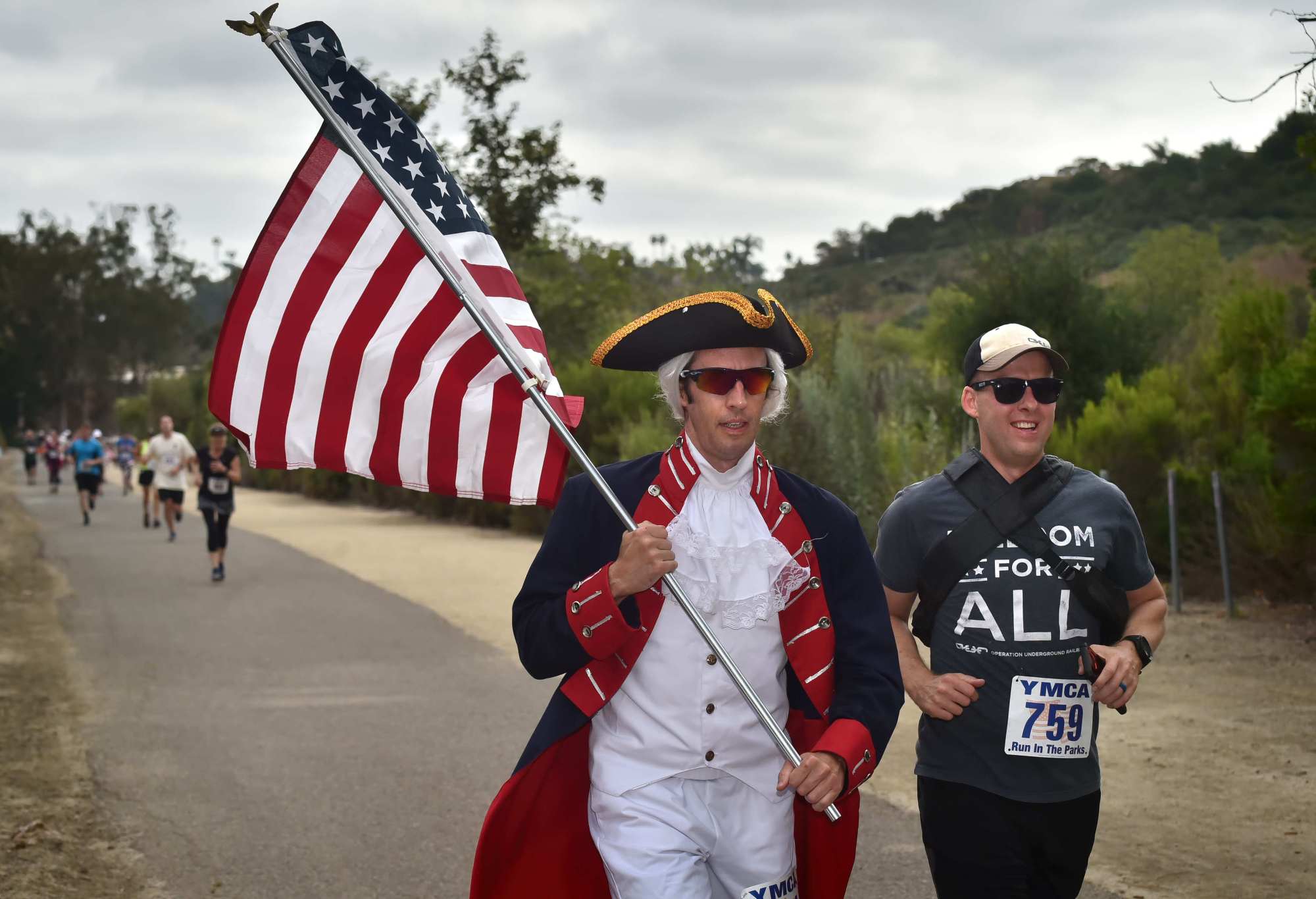 George Wilmington, left, runs with an American flag with Travis Weaver during the 41st Annual Fourth of July Run in the Parks, July 4, 2019 at Crown Valley Community Park. (Steven Georges, Contributing Photographer)