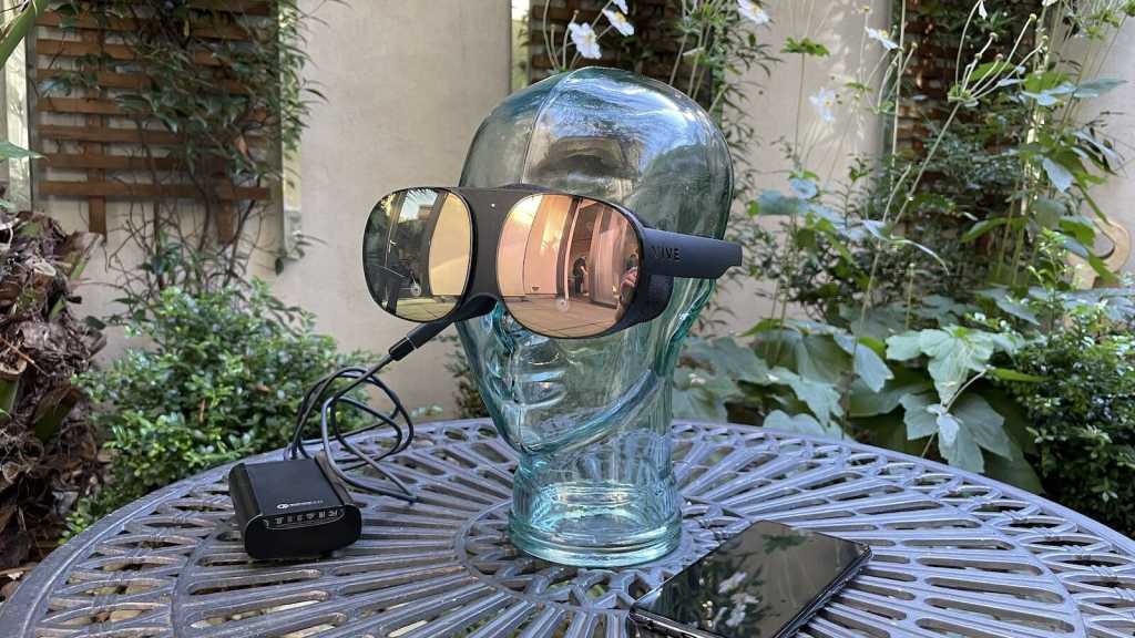 A shot of the Vive Flow on a glass head with a battery pack and smartphone on the table next to it