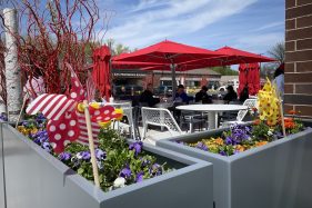 A patio surrounded by flowers, pinwheels and funky plantings. There are red umbrellas over the tables