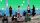 A photo of displaced Palestinians standing and seated on the side of a street with many of them using their mobile phones against a green grid background.