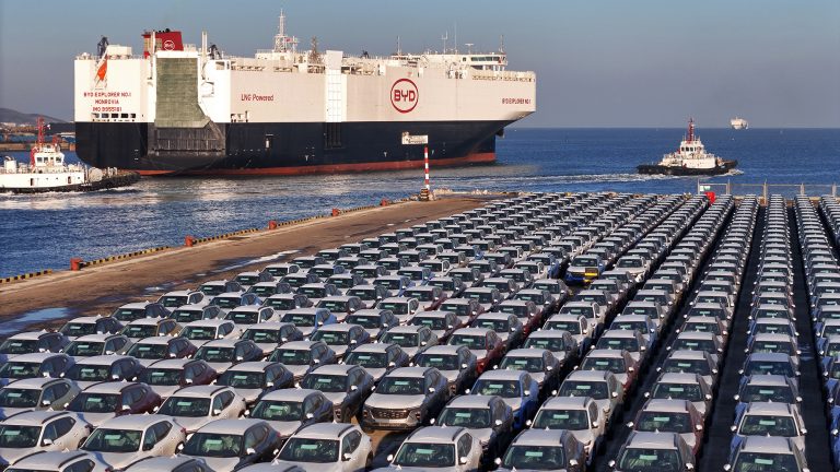 A photo showing rows of cars at a port in China with a large ship, with the label BYD on its side, setting off into the ocean.